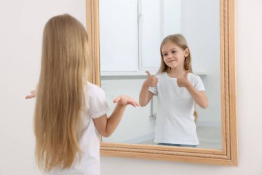 Aynanın önünde kafası karışmış küçük bir kız. Aynadaki yansıması parmaklarını içeride gösteriyor. Ruh Sağlığı