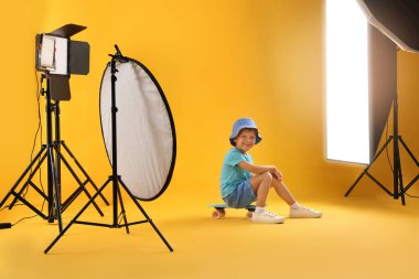 Sarı arka planda, profesyonel ekipmanlarla fotoğraf stüdyosunda para tahtasına sahip küçük bir çocuk.