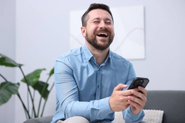 Akıllı telefonlu bir adam evdeki kanepede gülüyor.