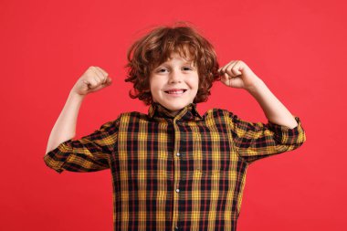 Kuvvet. Küçük mutlu çocuk kırmızı arka planda bisepslerini gösteriyor.