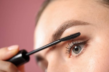 Genç bir kadın pembe arka planda kirpiklerine rimel sürüyor.