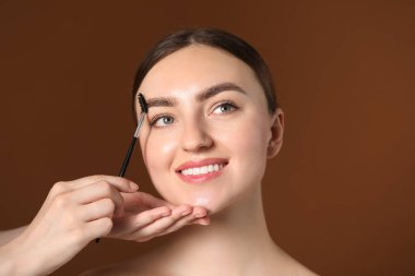 Kozmetikçi, kahverengi arka planda ürkütücü bir şekilde genç kadının kaşlarını fırçalıyor.