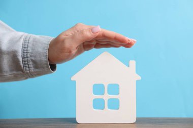 Ev güvenliği. Ahşap masadaki ev figürünü açık mavi arka plana karşı koruyan kadın, yakın plan.