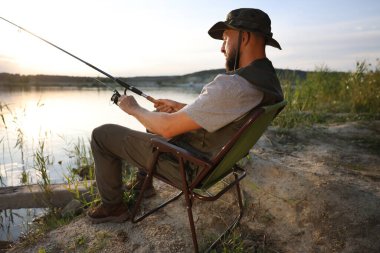 Yazın sandalyede oturan ve göl kenarında balık tutan oltalı balıkçı.