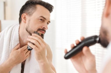 Yakışıklı adam evde aynanın yanında tıraş makinesiyle tıraş oluyor.