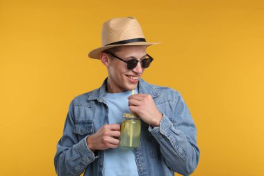 Sarı arka planda limonata kavanozu olan şapkalı ve güneş gözlüklü mutlu bir adam. Tazeleyici içecek.