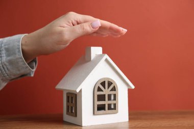 Ev güvenliği. Ahşap masadaki ev modelini kırmızı arka plana karşı koruyan kadın, yakın plan.