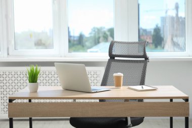 Ofisteki ahşap masada dizüstü bilgisayarı, kırtasiyesi, kahvesi ve çiçek dekoru olan şık bir iş yeri.