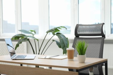 Ofisteki ahşap masada dizüstü bilgisayarı, kırtasiyesi, kahvesi ve çiçek dekoru olan şık bir iş yeri.