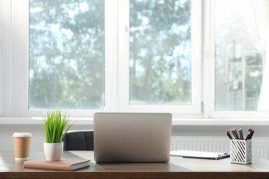 Ofisteki ahşap masada dizüstü bilgisayarı, kırtasiyesi, kahvesi ve çiçek dekoru olan şık bir iş yeri.