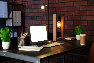 Laptop, paket kâğıt bardak, ev bitkileri ve tuğla duvarın yanındaki siyah masada kırtasiye malzemeleri.