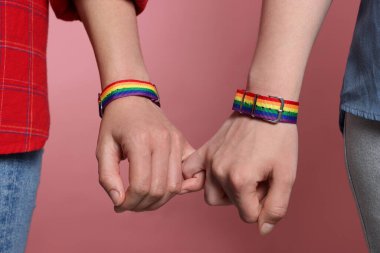 LGBT konsepti. Gökkuşağı bileklikli kadınlar pembe arka planda el ele tutuşuyorlar, yakın plan.
