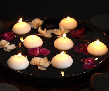 Yanan mumlar ve orkide çiçekleri tabakta süzülürken ve masadaki spa taşları siyah arkaplanda, yakın plan