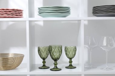 Beyaz duvarın yanında farklı bulaşık takımları ve gözlüklerle raf ünitesi.