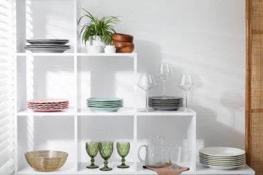 Beyaz duvarın yanında farklı bulaşık takımları, gözlük ve çiçek dekoru olan raf ünitesi.