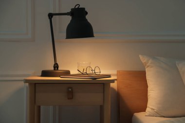 İçinde lamba, defter, bardaklar ve yatağın yanında bir bardak su olan komodin.