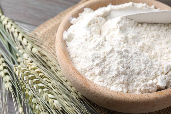 Flour in bowl and green wheat spikes on table, closeup