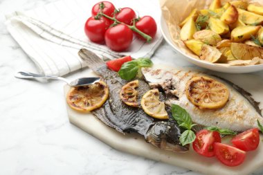 Beyaz mermer masada patates, domates, fesleğen ve limon dilimleriyle servis edilen kızarmış balık. Ev yapımı deniz ürünleri yemeği.