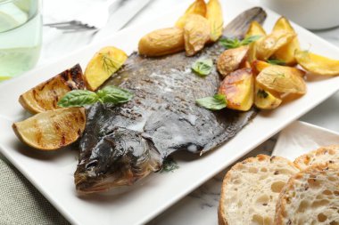 Beyaz mermer masada patates, fesleğen ve limon dilimleri ile servis edilen kızarmış balık, yakından. Ev yapımı deniz ürünleri yemeği.
