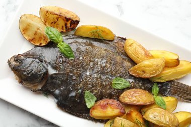 Beyaz mermer masada patates, fesleğen ve limon dilimleri ile servis edilen kızarmış balık, üst manzara. Ev yapımı deniz ürünleri yemeği.