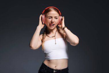 Şık bijouterie, kulaklıklar ve koyu gri arka planda poz veren oyuncu kadın.