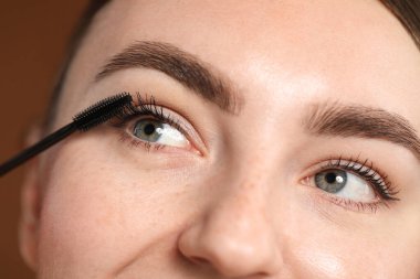 Genç bir kadın kahverengi arka planda kirpiklerine rimel sürüyor, yakın plan.