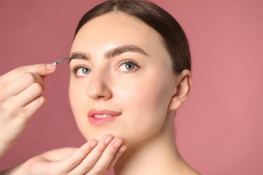 Kozmetikçi, pembe arka planda cımbızla genç bir kadının kaşını koparıyor.