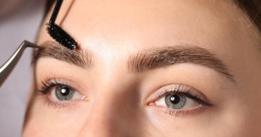 Açık gri arka planda, ürkütücü bir şekilde genç kadının kaşlarını yolmak ve fırçalamak. Yakın plan.