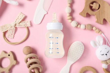 Açık pembe zemin üzerinde süt ve bebek aksesuarları olan bir şişe.
