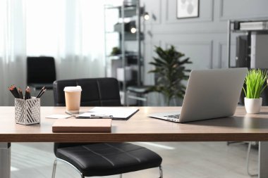 Ofisteki ahşap masada dizüstü bilgisayarı, kırtasiyesi, kahvesi ve çiçek dekoru olan şık bir iş yeri.