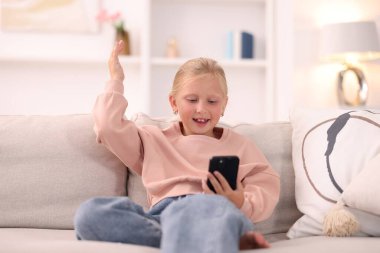 Akıllı telefonlu şirin bir kız evdeki kanepede gülüyor.