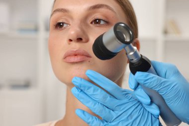 Doktor hastanede dermatoskopla genç kadının benini inceliyor.