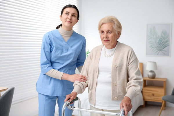 Nurse assisting senior woman with walking frame indoors. Home health care service