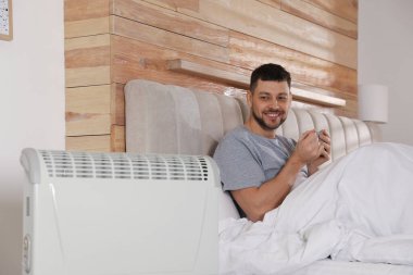 Evdeki elektrikli ısıtıcının yanındaki yatakta bir bardak sıcak içeceği olan adam.
