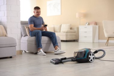 Evdeki koltukta akıllı telefonu olan adam, elektrik süpürgesine odaklan.