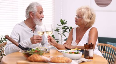 Yaşlı bir çift, akşam yemeği sırasında evde beyaz şarap bardaklarını tokuşturuyor.