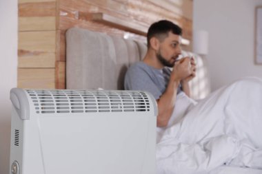 Evdeki yatakta bir bardak sıcak içeceği olan adam, elektrikli ısıtıcıya odaklan.