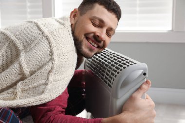 Evde elektrikli ısıtıcının yanında battaniyeli bir adam ısınıyor.