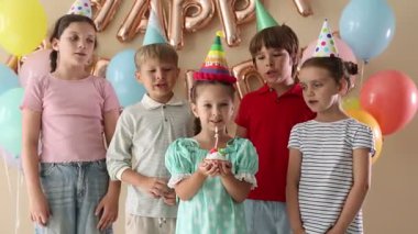 Doğum günü kutlaması. Tatlı küçük kız kekin üzerine mum üflüyor, arkadaşları ve bej arka planda şenlik dekoru.