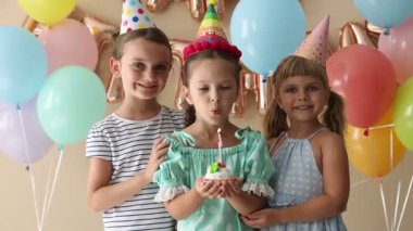 Doğum günü kutlaması. Tatlı küçük kız kekin üzerine mum üflüyor, arkadaşları ve bej arka planda şenlik dekoru.