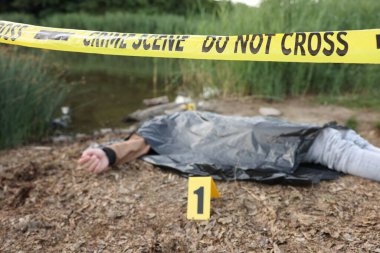 Adli tıp uzmanlığı. Cesedin bulunduğu suç mahalli, gölün yakınında işaret ve polis bandı, seçici bir odak noktası.