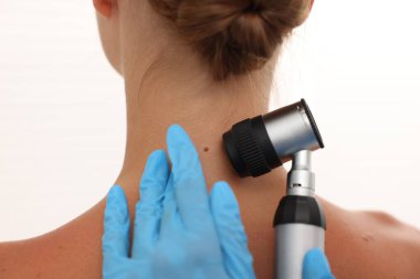 Doktor beyaz arka planda dermatoskopla genç kadının benini inceliyor.