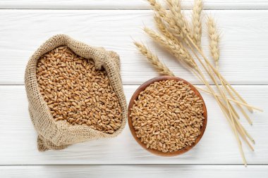 Kase içinde buğday taneleri, çuval bezi torbası ve beyaz ahşap masada çivili kazıklar, düz uzanmış.