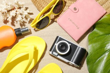 Yaz tatili zamanı. Kamera, pasaport, güneş kremi, yeşil yaprak ve kumsaldaki aksesuarlar.