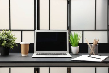 Laptop, paket kâğıt bardak, ev bitkileri ve kırtasiye malzemeleri evdeki siyah masada.