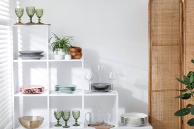 Beyaz duvarın yanında farklı bulaşık takımları, gözlük ve çiçek dekoru olan raf ünitesi.