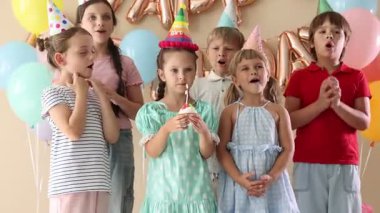 Doğum günü kutlaması. Tatlı küçük kız kekin üzerine mum üflüyor, arkadaşları ve bej arka planda şenlik dekoru.