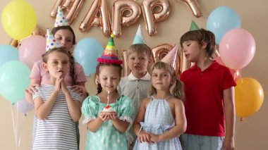 Doğum günü kutlaması. Tatlı küçük kız kekin üzerine mum üflüyor, arkadaşları ve bej arka planda şenlik dekoru.