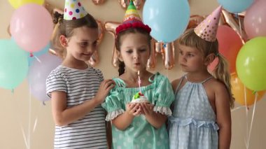 Doğum günü kutlaması. Tatlı küçük kız kekin üzerine mum üflüyor, arkadaşları ve bej arka planda şenlik dekoru.