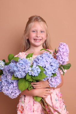 Bej arkaplanda bir buket hortensiya çiçeğiyle gülümseyen küçük kızın portresi.